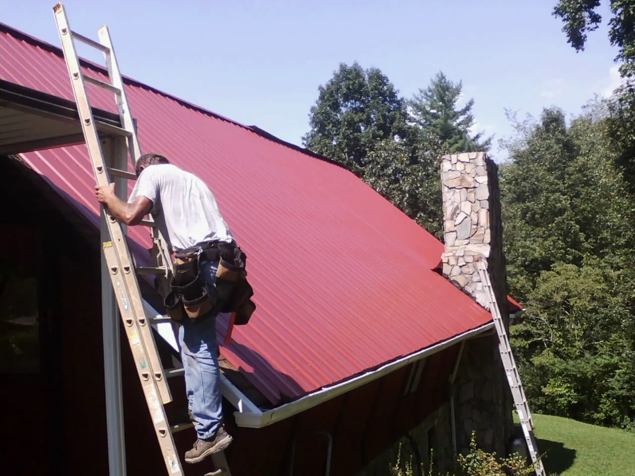 Roofer on a ladder beside a red metal roof and stone chimney — Soffit & Fascia Repair by Roofing Brothers in Kenosha